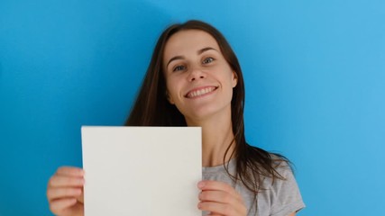 Portrait of beautiful smiling woman with long hair shaking her head, holding blank paper sheet isolated over blue background with copy space. People sincere emotions lifestyle. 