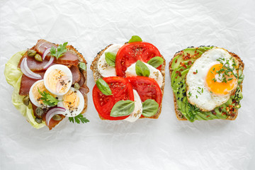 Variety of open sandwich,  wholemeal bread slices  with various toppings: smoked tuna, egg, lettuce, tomatoes, mozzarella, avocado, fried egg, fresh herbs on a white background, top view. 