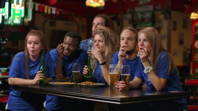 A multi-ethnic group of friends of fans in blue t-shirts will watch a match on TV in a bar, watch a tense exciting moment and celebrate a goal scored. Hugging, having fun, and making a glass of beer