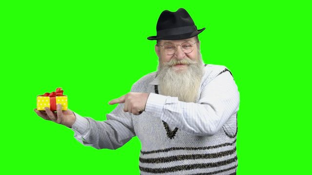 Positive senior man pointing at gift box. Elderly bearded man in hat showing gift box on green screen. Concept of discount.