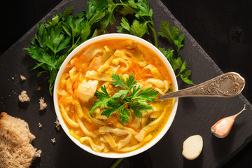 An overhead photo of a plate of chicken and noodles soup, shot from above on a dark rustic texture with a spoon, slices of bread, a celery branch, and garlic..