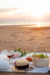 Picnic in the rays of the setting sun on the sand. On the plaid lies fruit basket, glasses, flowers and sweet pastries.