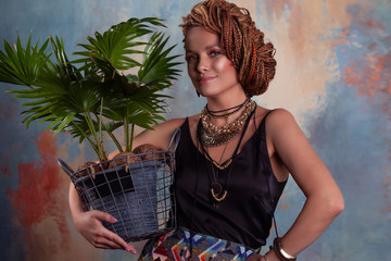 Southern flavor. A young tanned woman with afro braids smiles holding a big pot with a tropical plant.