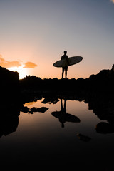 A male surfer at sunset comes with a surfboard over the cliffs of the ocean. Waiting for the wave. Silhouet of a sportsman guy in the rays of a summer warm sun.