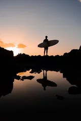 Ein männlicher Surfer kommt bei Sonnenuntergang mit einem Surfbrett über die Klippen des Ozeans. Warten auf die Welle. Silhouette eines Sportlers in den Strahlen einer warmen Sommersonne. © 072Y