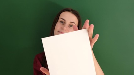 Closeup portrait of beautiful smiling female wearing red t-shirt, posing with white blank paper with copy space for your advertisement information and looking at camera, against green background.