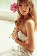Beautiful blonde woman with sensual lips posing in lightfull white room among peonies flowers