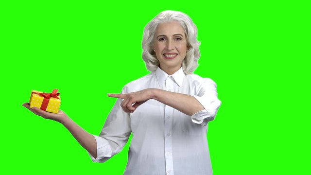 Smiling senior woman holding gift box on her palm. Pretty caucasian woman in elegant shirt pointing with finger on gift box. Green screen background.