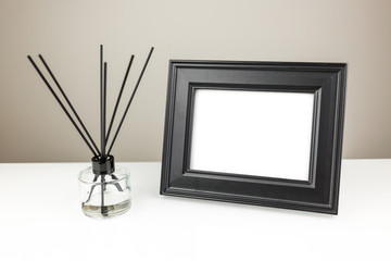 Aromatizer or perfume stick. Aromatic air freshener in a transparent glass bottle with black reeds on a white table and black desk photo frame with a blank space on a white table