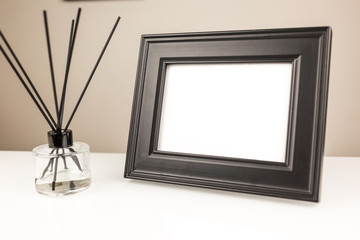 Aromatizer or perfume stick. Aromatic air freshener in a transparent glass bottle with black reeds on a white table and black desk photo frame with a blank space on a white table