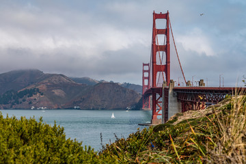 Golden Gate Bridge on a sunny day with some grass and bushes in the foreground and a sailboat to the left of the bridge