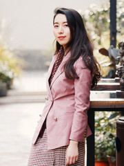 Beautiful Chinese young woman in pink suit dress posing and smiling at camera with blur outdoor cafe background, side view, Outdoor fashion portrait of glamour stylish lady.