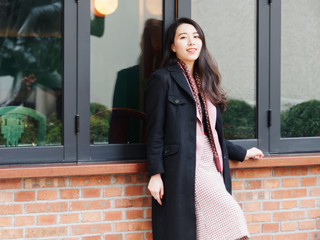 Beautiful Chinese young woman in black coat and pink suit dress posing and smiling at camera in front of glass windows and brick wall, front view, Outdoor fashion portrait of glamour stylish lady.