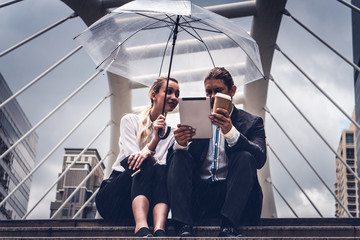 two businesspeople with umbrella watching tablet and having business discussion in rainy season