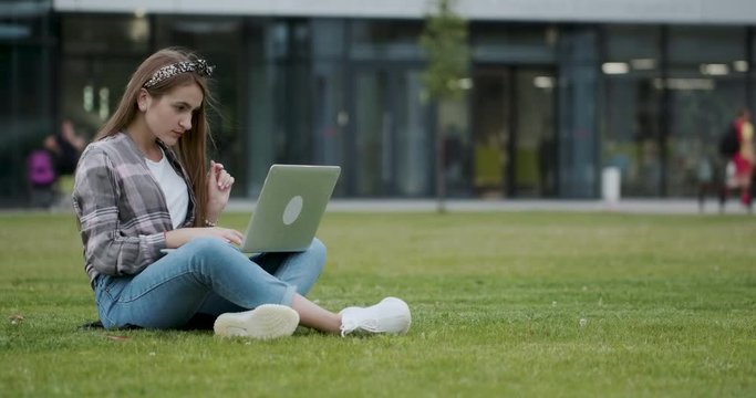 Girl using a laptop computer to look up information on the internet. A pretty female college student sits in shade on grass near public garden.