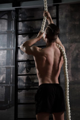Fototapeta na wymiar Muscular guy with a naked torso in sports shorts holding on to a rope while standing in the gym, frame from the back.
