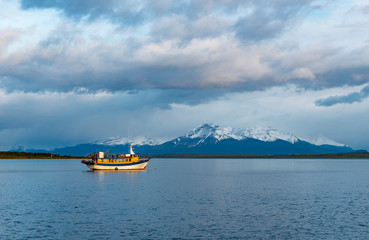 Landscape of the Last Hope Sound at sunrise with a fishing boat in the foreground and snowcapped Andes peaks in the background, Puerto Natales, Patagonia, Chile.