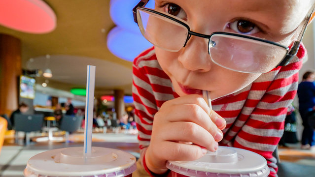 Cute little blond child in glass drinks carbonated drink and eats burger in fast food cafe. Concept of fast food. Shallow focus, blur.
