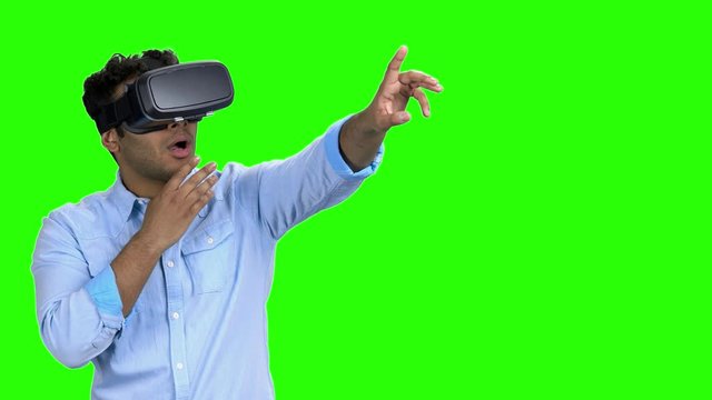 Amazed young man using virtual reality glasses. Shocked indian man wearing VR googles pointing at someting with index finger. Green screen background.