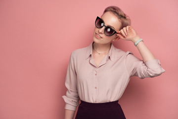 A girl with short pink hair in a blouse and skirt, smiling, looking at the camera in sunglasses, standing on a pink background.