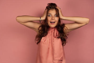 Fototapeta premium A dark-haired girl in a pink dress stands on a pink background with her hands on her head, her mouth wide open.