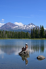 Young woman sitting on rock enjoys view of Middle and North Sisters volcanoes reflected in Scott Lake, Oregon on a calm sunny summer afternoon.
