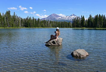 Obraz na płótnie Canvas Young woman sitting on rock enjoys view of Middle and North Sisters volcanoes reflected in Scott Lake, Oregon on a calm sunny summer afternoon.