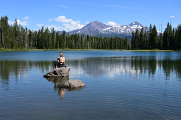 Young woman sitting on rock enjoys view of Middle and North Sisters volcanoes reflected in Scott Lake, Oregon on a calm sunny summer afternoon.