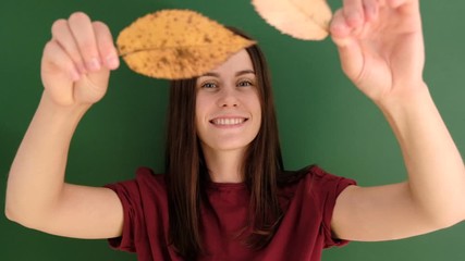 Cheerful young girl with long hair wearing red t-shirt, having fun playing with yellow autumn leaves and looking at camera, against green background. People and lifestyle.