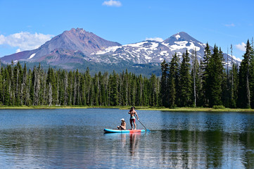 Fototapeta na wymiar Young couple paddle boarding on Scott Lake, Oregon, with Middle and North Sisters volcanoes in the background on a calm sunny summer afternoon.