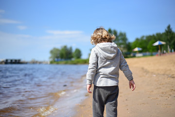 Blond kid walking on the beach in light clothes. Warm summer day. A lonely boy on the river Bank. Emotions. Walk with children. rear view.