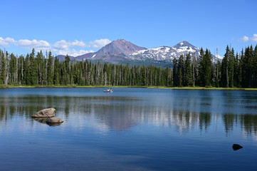 Two young women on standup paddle board on Scott Lake, Oregon with Middle and North Sisters volcanoes in background.