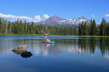 Two young women on standup paddle board on Scott Lake, Oregon with Middle and North Sisters...