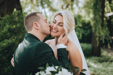 Beautiful newlyweds hug and kiss on the nature in greenery, in the park. Wedding portrait of a stylish groom in a suit and a smiling blonde bride close-up. Concept and photography.