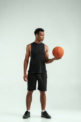 Naklejka premium Ready to play. Full-length of confident young african man carrying a basketball ball and looking at it while standing in studio against grey background