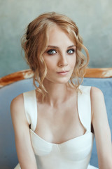Beautiful slender blonde girl sitting on the sofa in a long white dress. Portrait of a woman with a flower in hand. Perfect hairstyle and cosmetics of the bride, new collection of wedding dresses