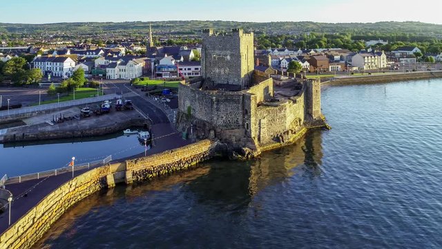 Belfast Lough with Medieval Norman Castle in Carrickfergus near Belfast in sunrise light. Aerial 4K flyby video with marina, breakwater, yachts, parking, town and far view of Belfast in the background