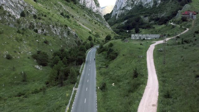 A tracking aerial drone shot, following a cyclist on a meandering road in between a vast gorge and far mountains, in Coltesti, Romania. Trees line the road, a couple of houses populate the shot.