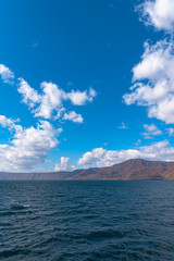 Beautiful autumn foliage scenery landscapes. Fall is full of magnificent colors. View from Lake Towada sightseeing Cruise ship. Clear blue sky, water, white cloud, sunny day background. Aomori, Japan