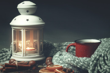Lantern with a burning candle, spices and a red mug with hot coffee on a snowy wooden table