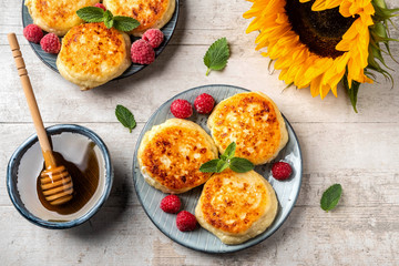 Cottage cheese pancakes with raspberries and mint leaves on a plate, wooden light background, top view, copy space. Food flat lay with sunflowers, pancakes and honey.
