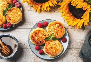 Cottage cheese pancakes with raspberries and mint leaves on a plate, wooden light background, top view, copy space. Food flat lay with sunflowers, pancakes and honey.