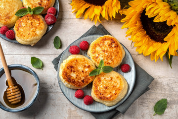 Cottage cheese pancakes with raspberries and mint leaves on a plate, wooden light background, top view, copy space. Food flat lay with sunflowers, pancakes and honey.