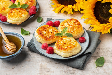 Cottage cheese pancakes with raspberries and mint leaves on a plate, sunflowers, and honey, light background.