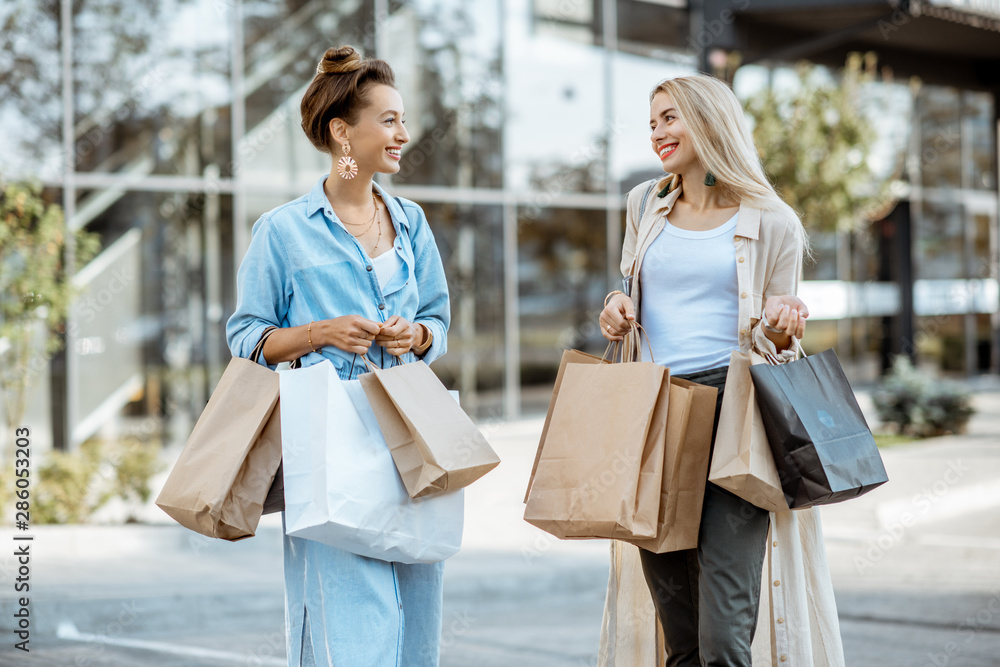 Wall mural Two happy girlfriends feeling satisfied with purchases, standing together with shopping bags in front of the shopping mall outdoors