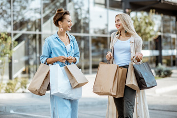 Two happy girlfriends feeling satisfied with purchases, standing together with shopping bags in...