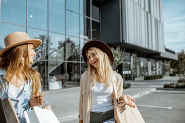 Two happy girlfriends walking with shopping bags in front of the shopping mall, feeling satisfied with new purchases