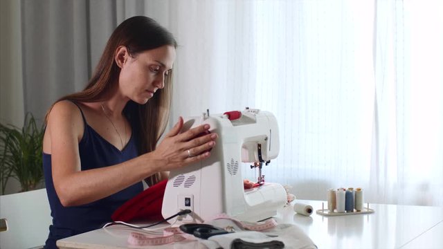 Young woman tailor is sewing red cloth on sewing machine. Seamstress is working at home sitting at table in her workplace. Self-employed people concept. Custom tailoring clothes for sale.