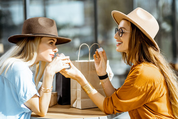 Two stylish girlfriends sitting at the modern cafe terrace outdoors, looking on a new purchases...