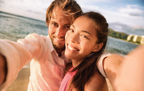 Happy smiling biracial couple taking selfie photo with camera phone. Caucasian man holding smartphone on beach vacation holiday sunset.
